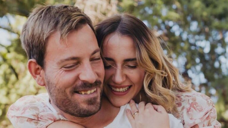 Couple smiling outdoors as the woman embraces the man from behind, both looking happy.