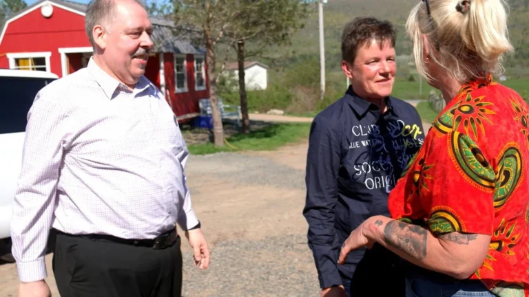 Three adults standing on a gravel driveway, chatting outdoors with a red barn and trees in the background. From left to right: a man in a light check shirt, a woman in a dark jacket, and a woman in a bright red patterned shirt showing tattoos on her forearm.