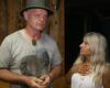 Man in a green hat speaks to a woman with long gray hair in a wood-framed workshop.
