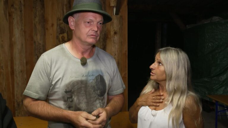 Man in a green hat speaks to a woman with long gray hair in a wood-framed workshop.