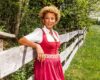 Woman in a red dress with a white blouse leaning on a wooden fence in a rural field, smiling at the camera.
