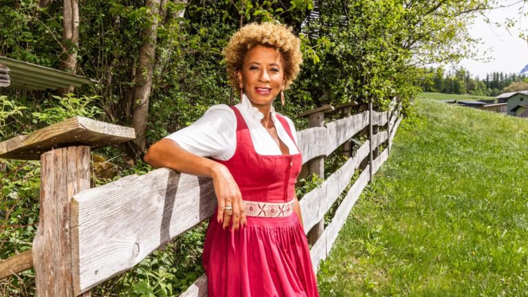 Woman in a red dress with a white blouse leaning on a wooden fence in a rural field, smiling at the camera.