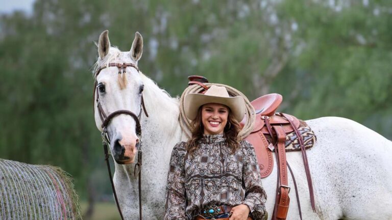 Smiling woman in a paisley blouse and cowboy hat standing beside a saddled white horse outdoors.