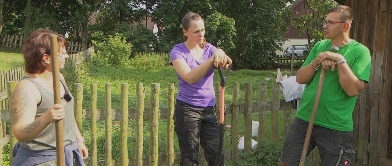 Three adults outdoors with shovels near a wooden fence, likely gardening or digging together.