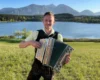 Man wearing a light green shirt plays a large accordion in a grassy field with a lake and mountains in the background.