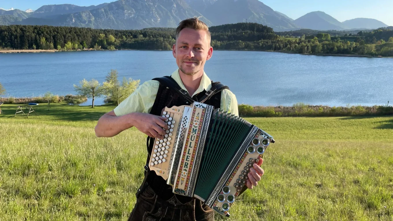 Man wearing a light green shirt plays a large accordion in a grassy field with a lake and mountains in the background.