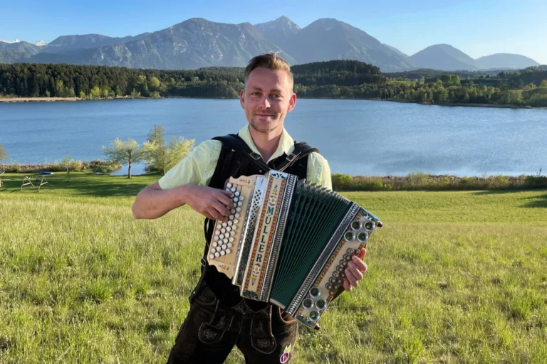 Man wearing a light green shirt plays a large accordion in a grassy field with a lake and mountains in the background.
