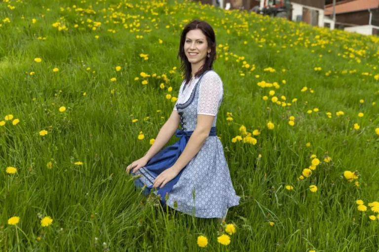 Woman in a blue dirndl sitting in a field of yellow dandelions, smiling at the camera.