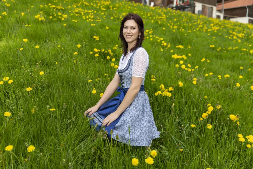 Woman in a blue dirndl sitting in a field of yellow dandelions, smiling at the camera.
