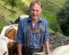 Smiling man in blue plaid shirt with leather suspenders standing among cows on a hillside.