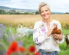 Smiling woman with short blonde hair holding a brown chicken in a sunny countryside field, goats in the background.
