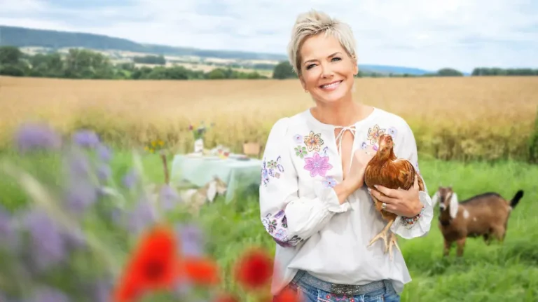 Smiling woman with short blonde hair holding a brown chicken in a sunny countryside field, goats in the background.