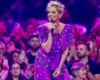 Female singer with short blond hair in a purple bejeweled dress performs on stage to a cheering crowd.