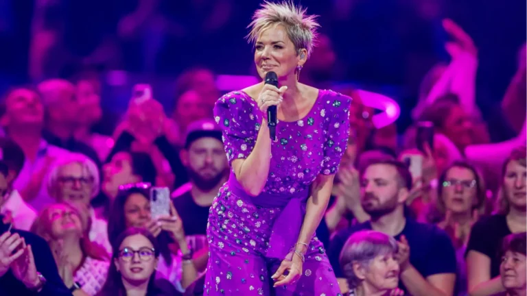 Female singer with short blond hair in a purple bejeweled dress performs on stage to a cheering crowd.