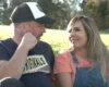 A man and woman sit close together in a sunlit field, smiling at each other.