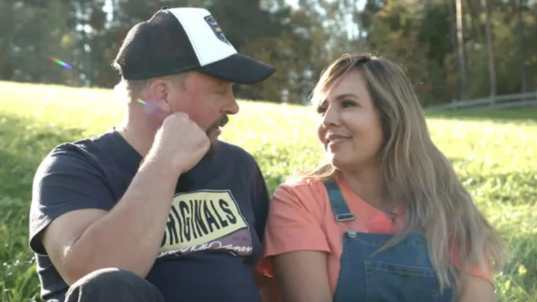 A man and woman sit close together in a sunlit field, smiling at each other.