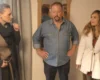 Three adults posing indoors: a woman in a dark coat and scarf left, a man in a denim shirt center, and a woman in a white coat right.