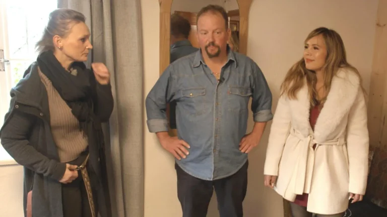 Three adults posing indoors: a woman in a dark coat and scarf left, a man in a denim shirt center, and a woman in a white coat right.