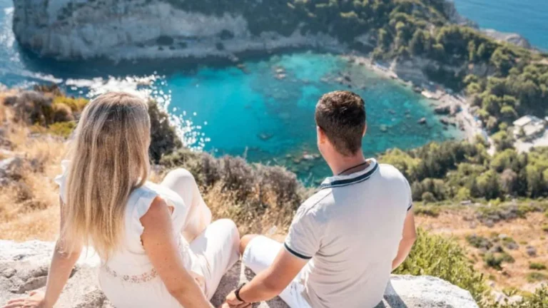 Couple sitting on a rocky ledge overlooking a turquoise bay and beach below, facing away from the camera.