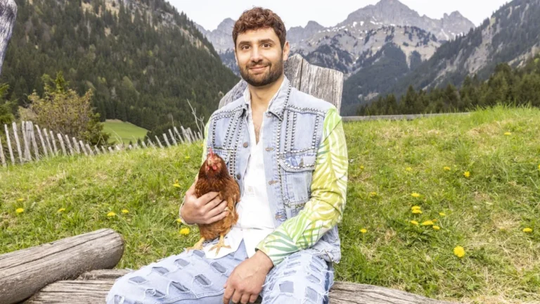 Man sits on a log in a meadow with mountains, holding a chicken.