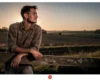 Man leaning on a weathered wooden fence at sunset in a rural field, gazing to the right.