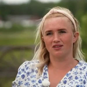 Woman with blonde hair wearing a pink headband and a white-blue floral blouse, speaking outdoors with a lapel mic visible on her chest.