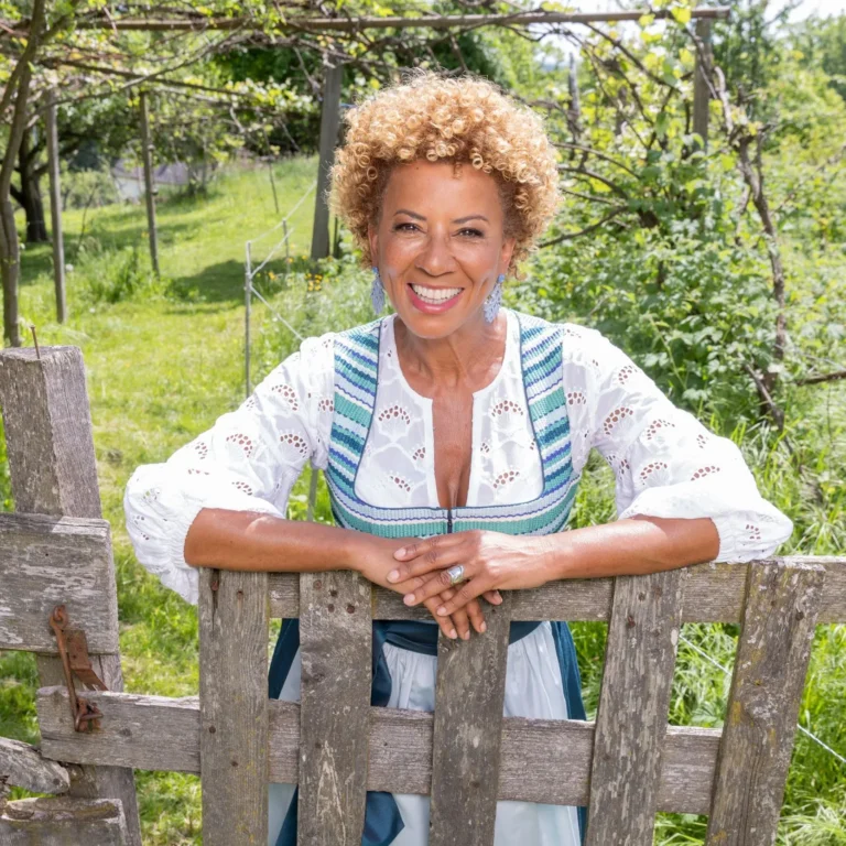 Smiling woman with curly blonde hair leaning on a wooden fence in a sunny garden.