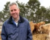 Smiling man in a blue jacket standing among cows in a farmyard with trees in the background.