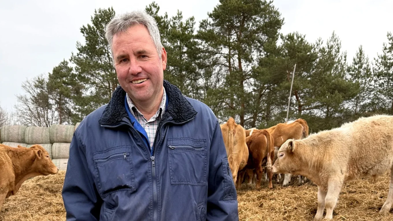 Smiling man in a blue jacket standing among cows in a farmyard with trees in the background.