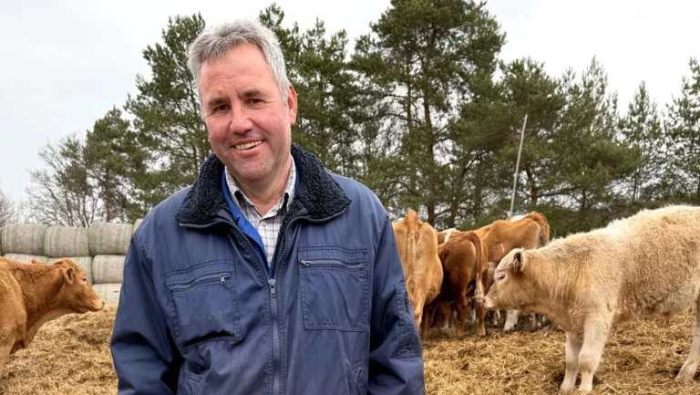 Smiling man in a blue jacket standing among cows in a farmyard with trees in the background.