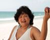 Smiling woman at the beach wearing a white sleeveless top, holding a light-colored ice cream cone visible at the edge of the frame.