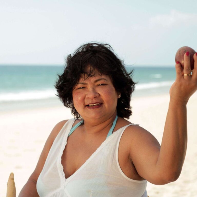Smiling woman at the beach wearing a white sleeveless top, holding a light-colored ice cream cone visible at the edge of the frame.