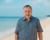 Middle-aged man wearing a blue patterned shirt stands on a sunny beach with the sea behind him.