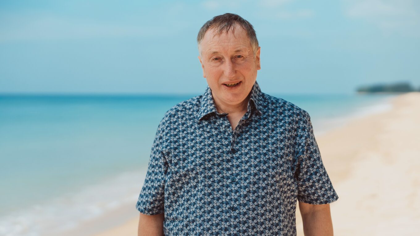 Middle-aged man wearing a blue patterned shirt stands on a sunny beach with the sea behind him.