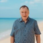 Middle-aged man wearing a blue patterned shirt stands on a sunny beach with the sea behind him.