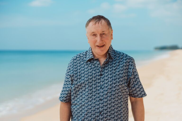 Middle-aged man wearing a blue patterned shirt stands on a sunny beach with the sea behind him.