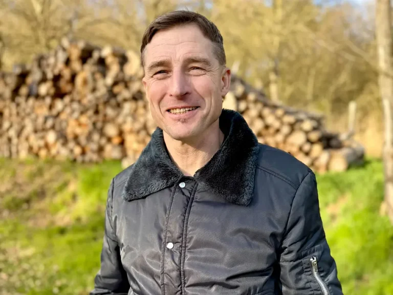 Smiling man in a gray jacket with a black fur collar outdoors, with a stack of cut logs in the background.