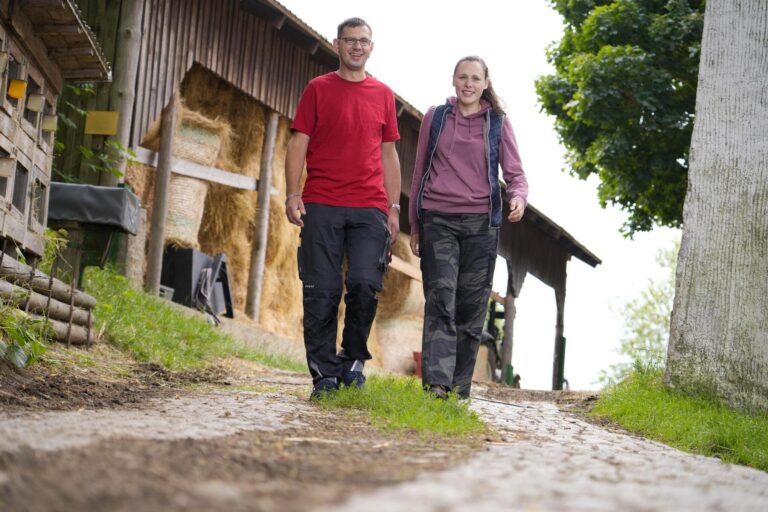 Two people walking along a rural farm path beside a wooden barn and hay bales, smiling at the camera.