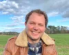 Smiling man in a shearling-lined brown jacket in a grassy field under a cloudy blue sky.