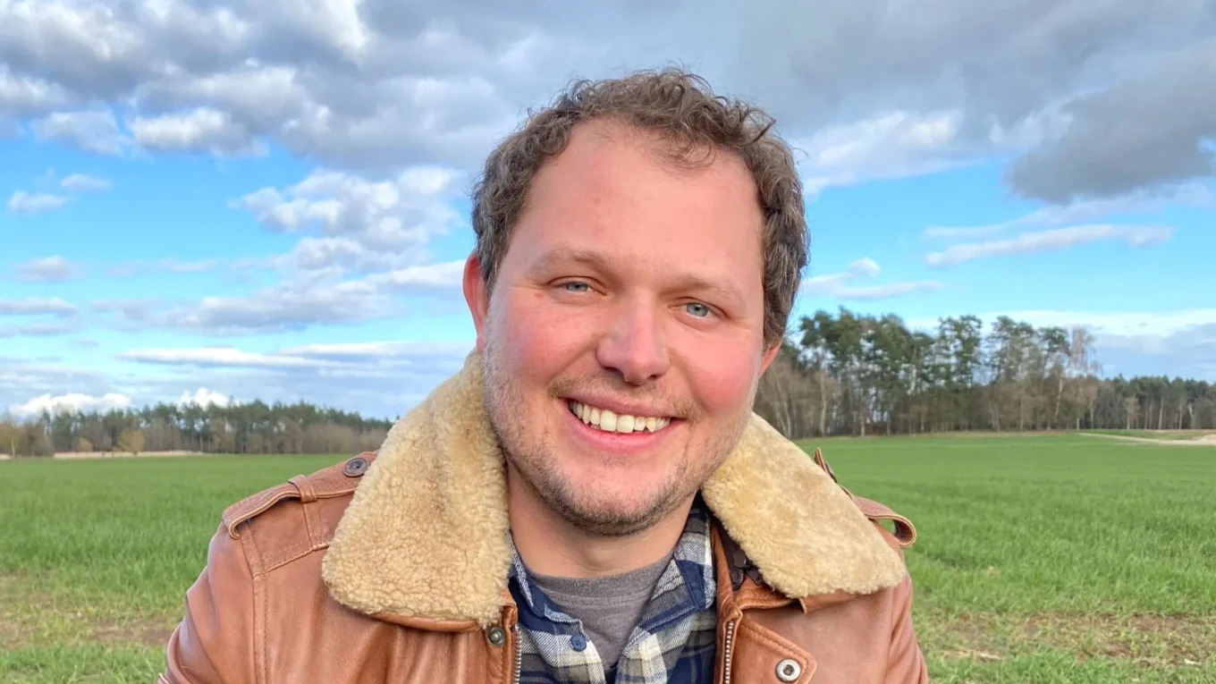 Smiling man in a shearling-lined brown jacket in a grassy field under a cloudy blue sky.