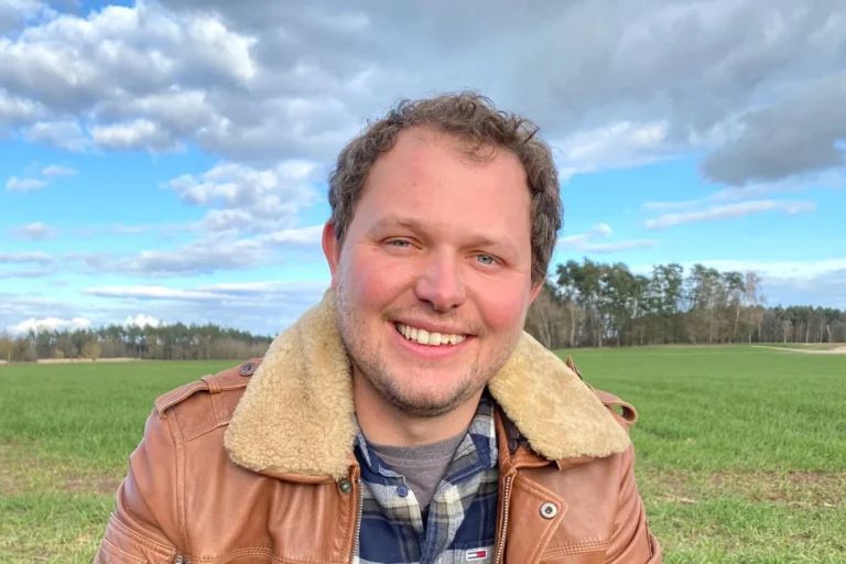 Smiling man in a shearling-lined brown jacket in a grassy field under a cloudy blue sky.